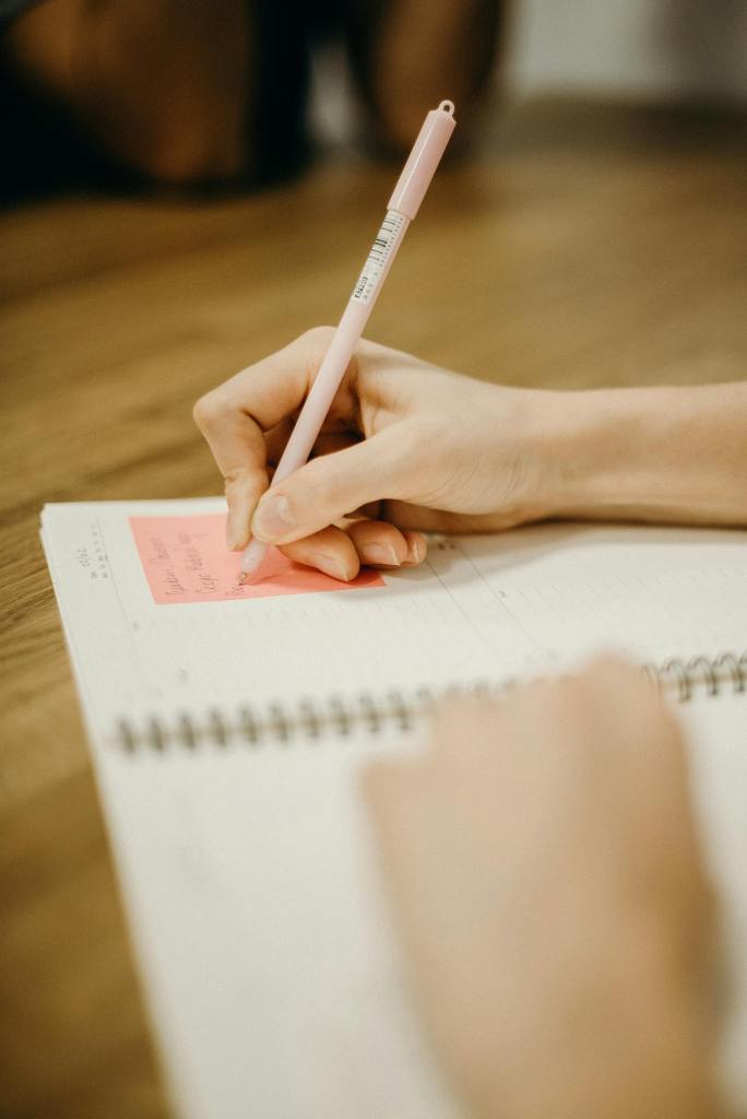 Close-up of a person writing in a notebook with a pink pen on a wooden desk.
